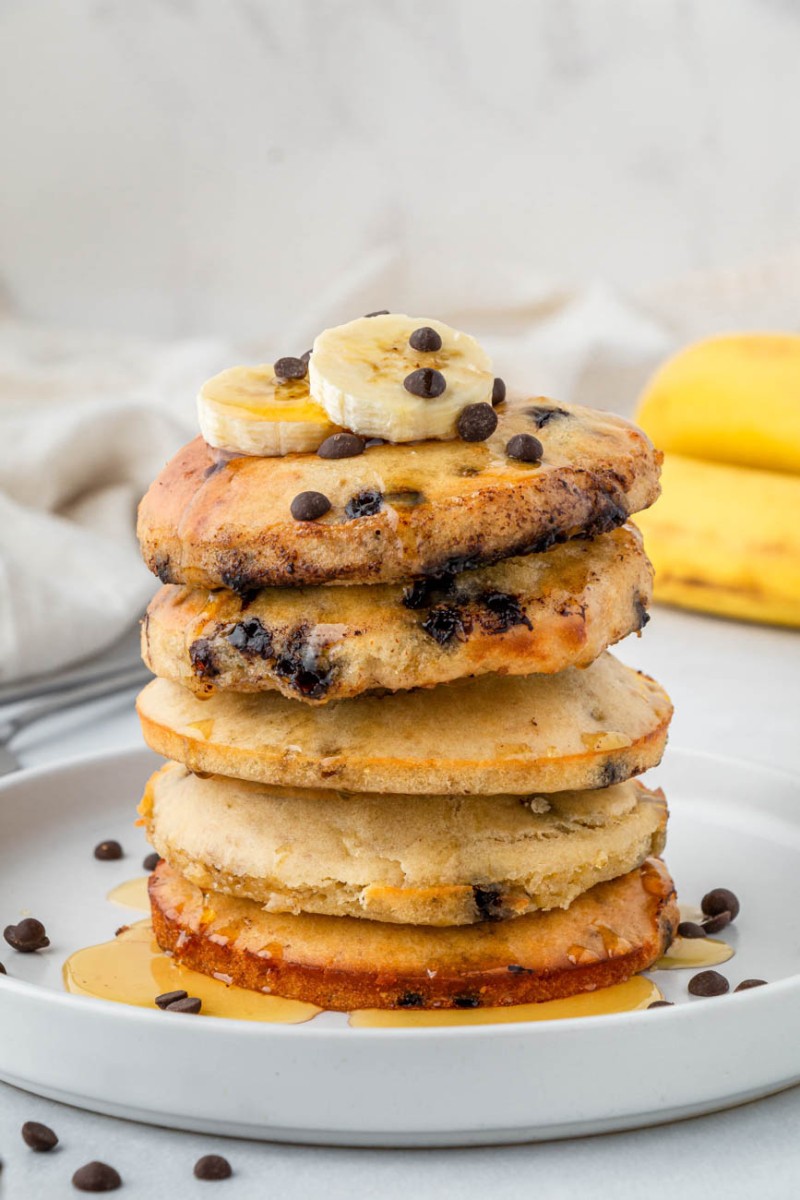 air fryer pancakes stacked on a white plate with chocolate chips, bananas, and syrup in the background