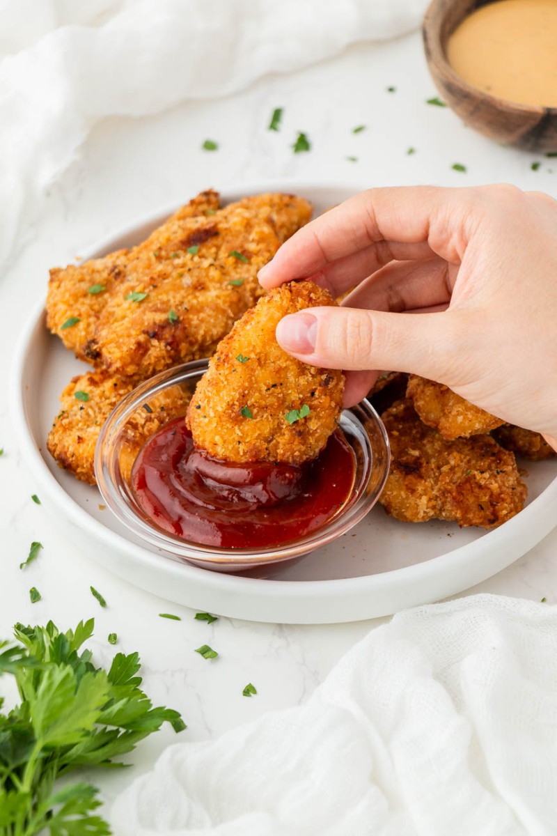 a hand dipping a chicken nugget in ketchup