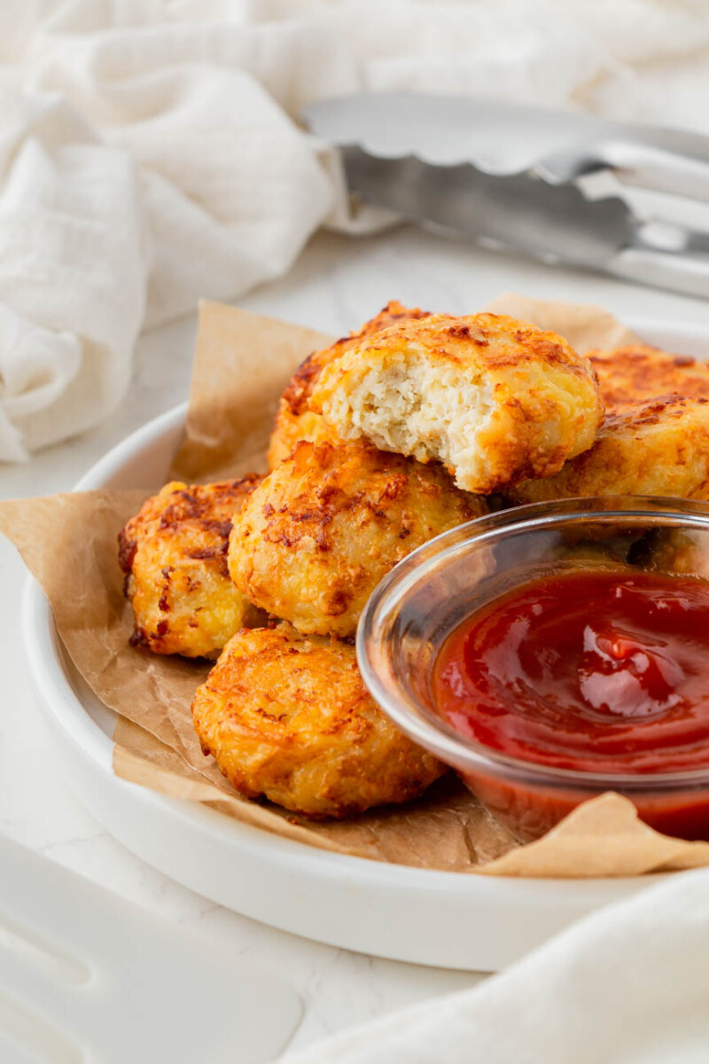 air fryer chicken nuggets on a plate, a bite taken out of one and a small bowl of ketchup