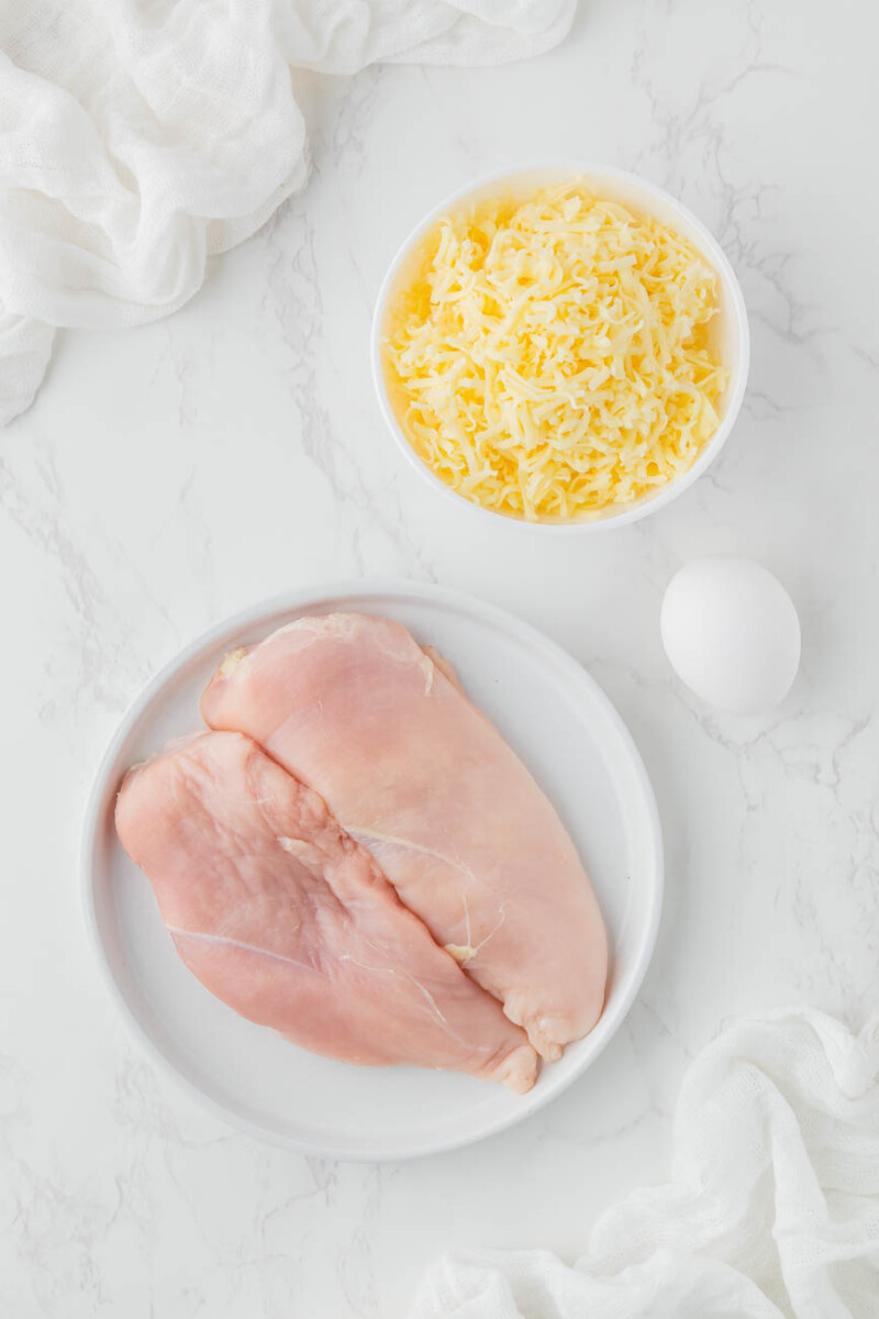 two chicken breasts on a plate, shredded cheese in a bowl, and an egg