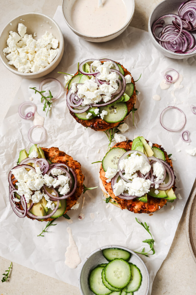 three open faced salmon burgers with cucumbers, red onion and feta cheese in small bowls on the side