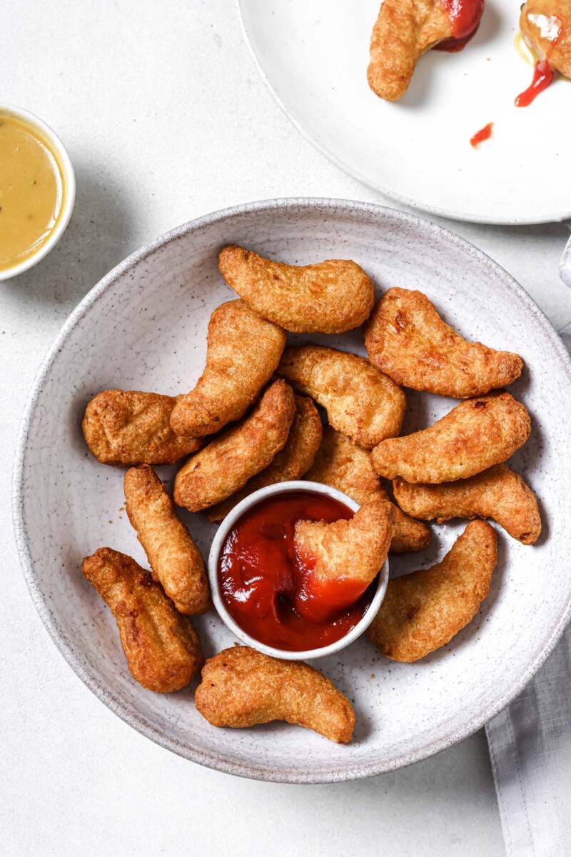 air fryer frozen chicken tenders in a white bowl with a small cup of ketchup and a piece of chicken dipped in it