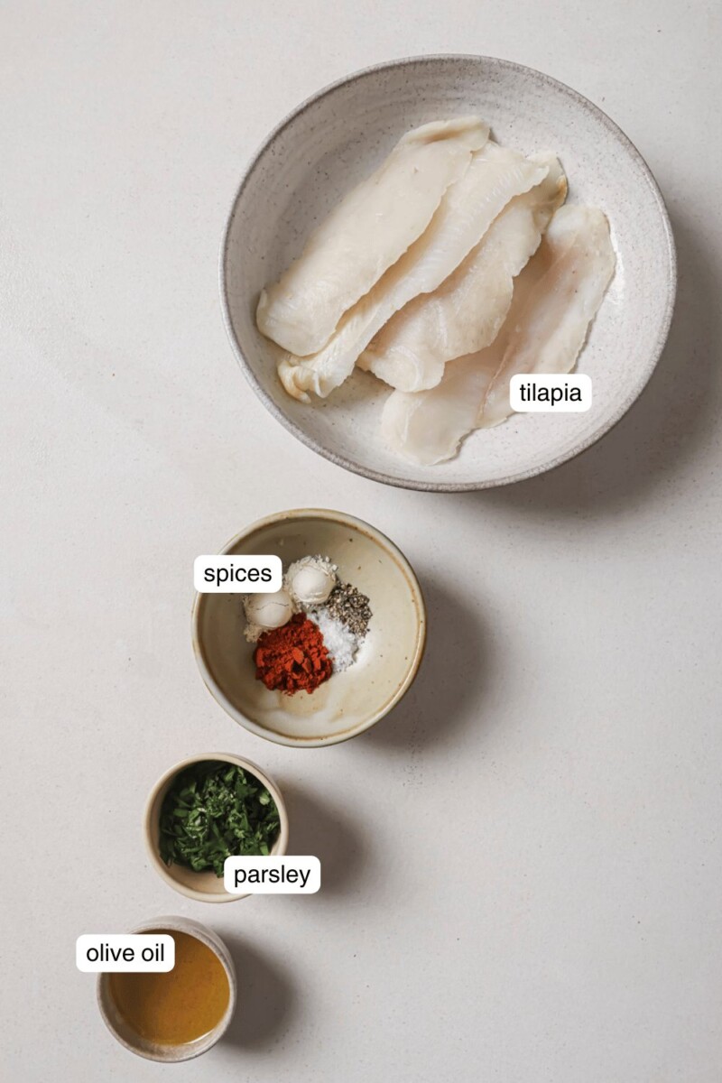 tilapia in a white bowl, spices, olive oil, and parsley in small ingredient bowls