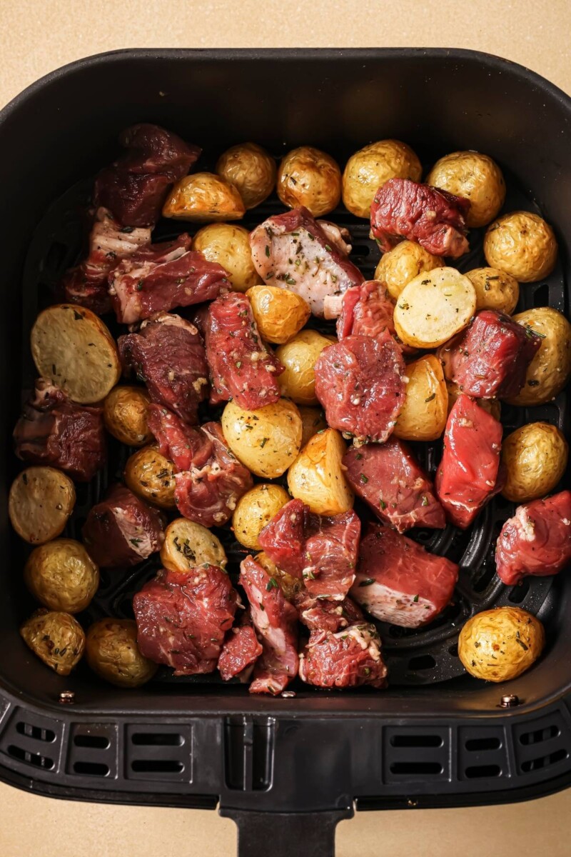 Uncooked potatoes and steak in an air fryer basket.