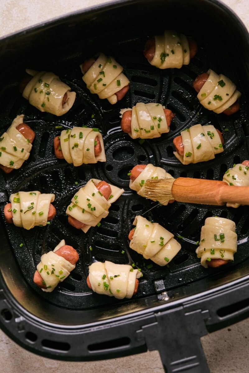 Brushing uncooked pigs in a blanket in the air fryer basket with melted butter.